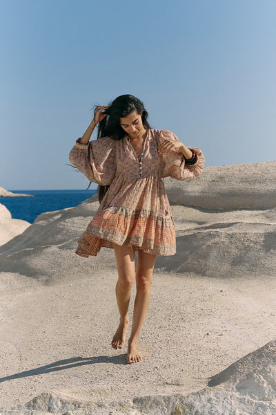 Woman on coastal rock with ocean and blue sky wearing the Daughters of India Bhoomi Mini Dress in Sunset, showing the tiered boho silhouette with flowing hem