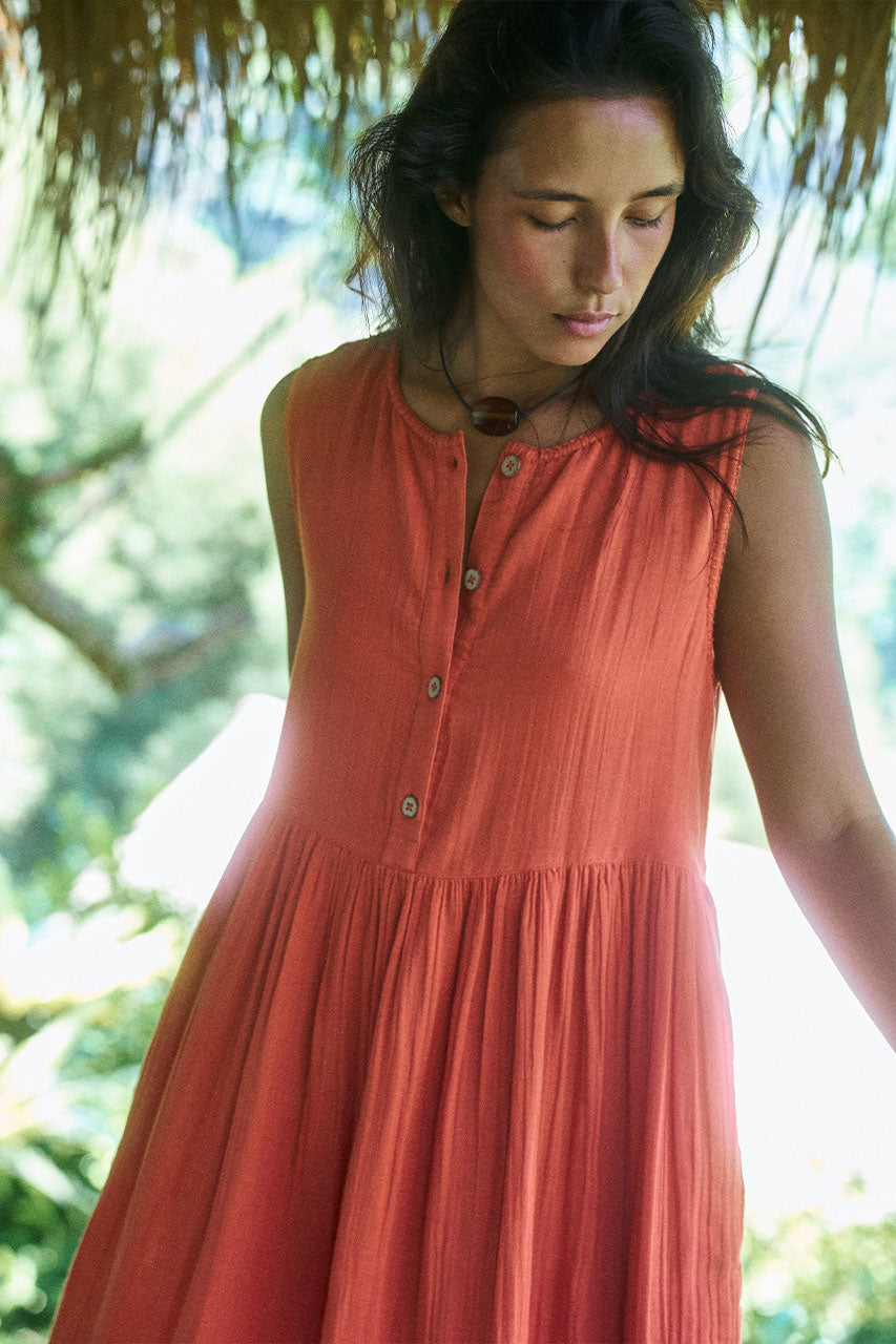 Nadi Dress Terracotta — upper body front view looking down, round neckline and wooden button front detail, terracotta double gauze cotton midi dress, tropical thatched roof setting | Daughters of India
