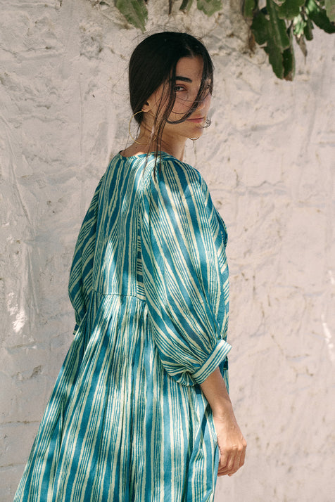 Woman standing against a white textured wall wearing the Daughters of India Kyra Maxi Dress in Spearmint, showing the green and white striped cotton fabric