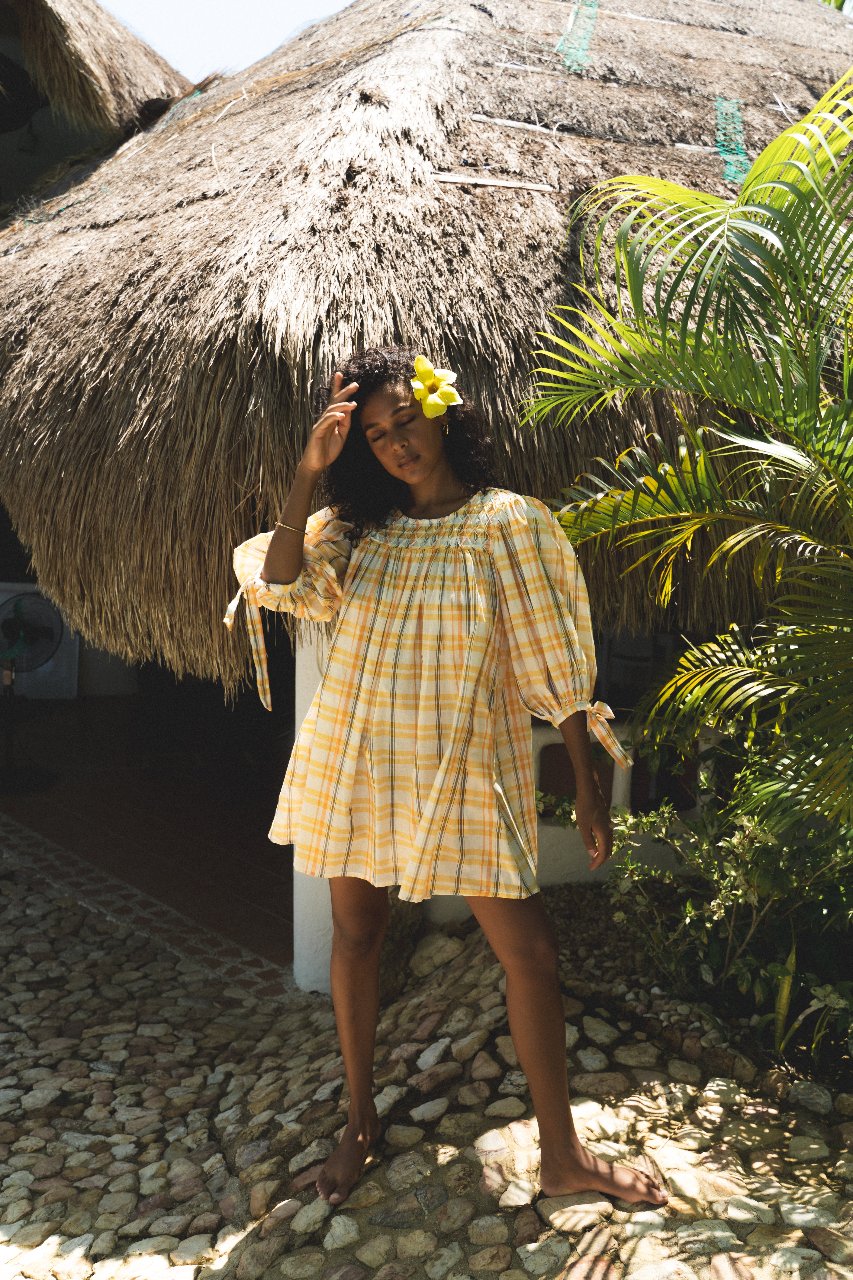 Freya Dress Honeycomb Plaid — full length outdoor on cobblestone, yellow flower in hair by thatched wall, cuff ties visible, handloom honeycomb plaid mini, ethical slow fashion artisan dress | Daughters of India